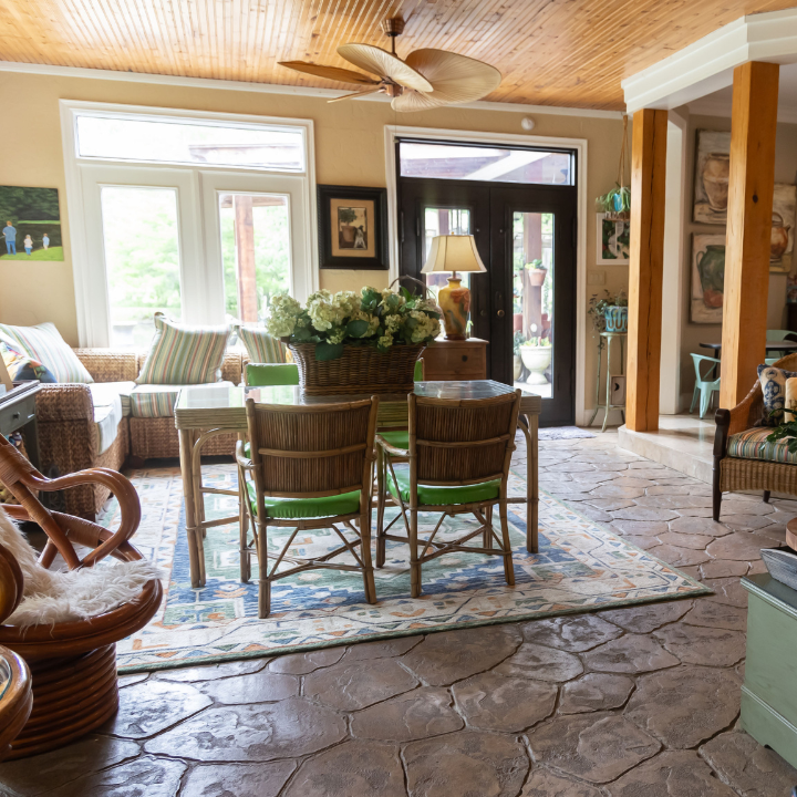 A sunroom features a dining set with wicker chairs, a stone floor, a wooden ceiling, and a large sectional sofa.
