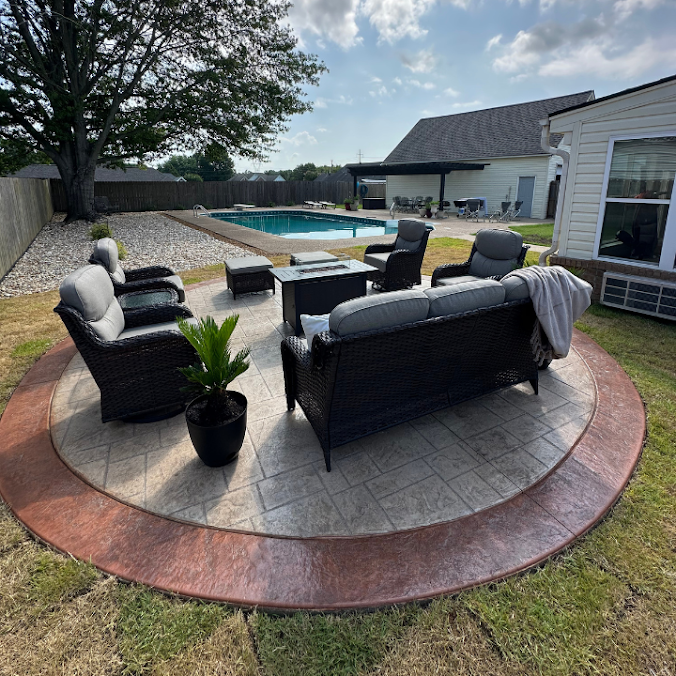 A circular stamped concrete patio with dark outdoor wicker furniture around a fire pit, located near a pool and house.
