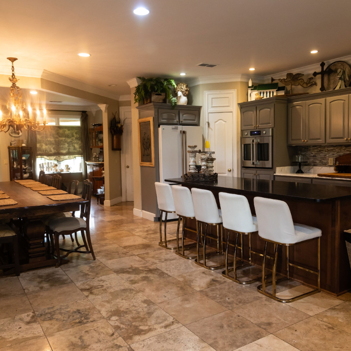 A spacious kitchen with a dark wood island, white bar stools, grey cabinets, and an adjacent dining area with stone floors.