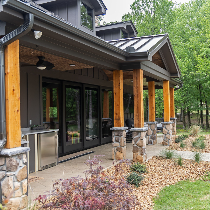 A modern home patio with dark gray siding, wooden support pillars on stone bases, metal roofing, and outdoor appliances.