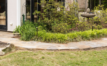 A stone garden path curves past lush green plants, a birdhouse, and a fountain in front of a house.