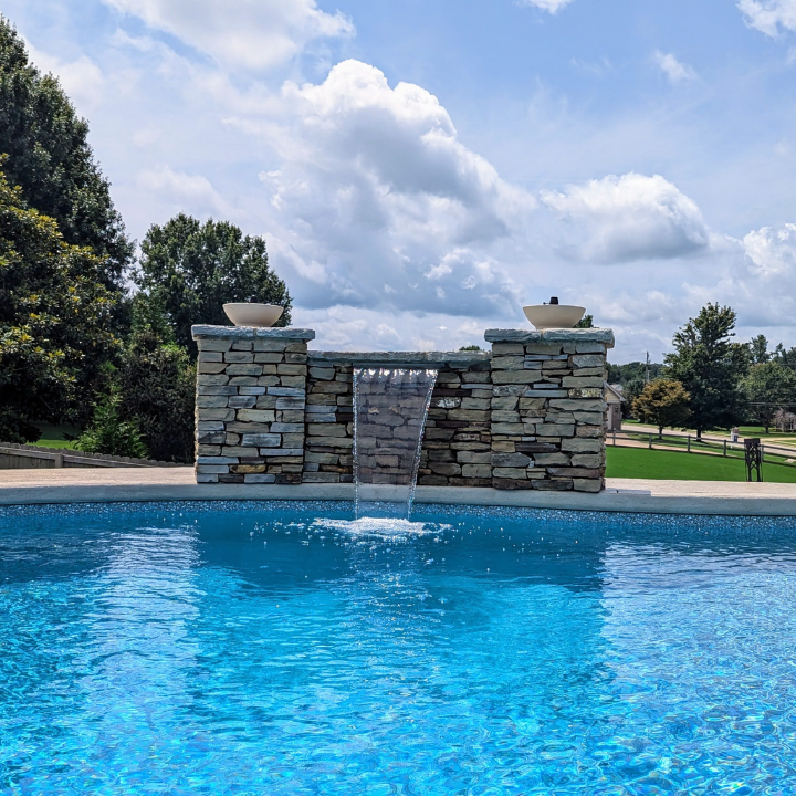 A stone waterfall feature built into a backyard swimming pool under a bright blue, cloudy sky.