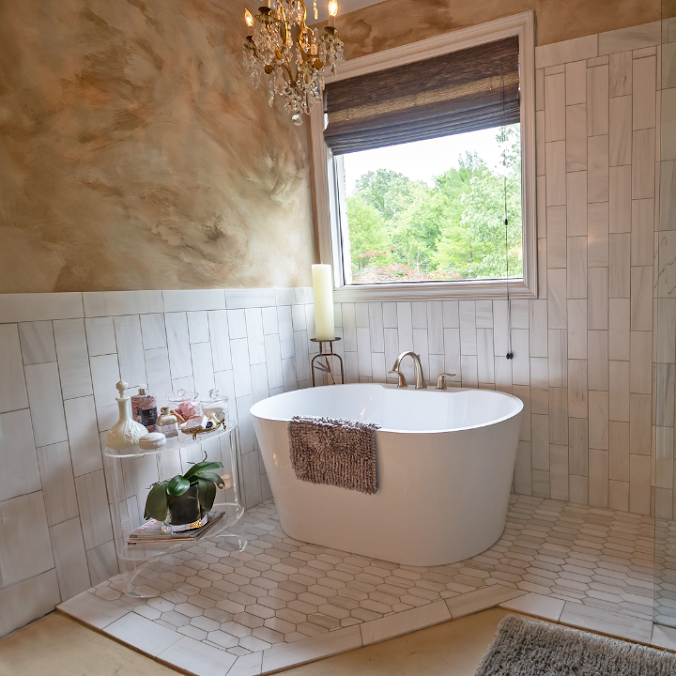 A freestanding white bathtub sits on a tiled floor near a window, accented by a chandelier and a small acrylic side table.
