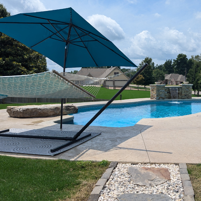 A hammock under a blue patio umbrella on a pool deck, with a swimming pool and stone water feature in the background.