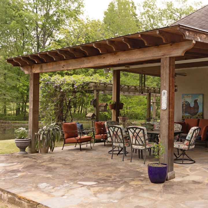 A flagstone patio under a wooden pergola, featuring outdoor chairs and a dining set, overlooking a lush, wooded backyard.