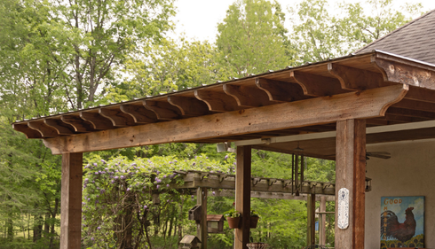 A rustic wooden porch overhang with decorative cut rafters, supported by timber posts, set against a backdrop of trees.