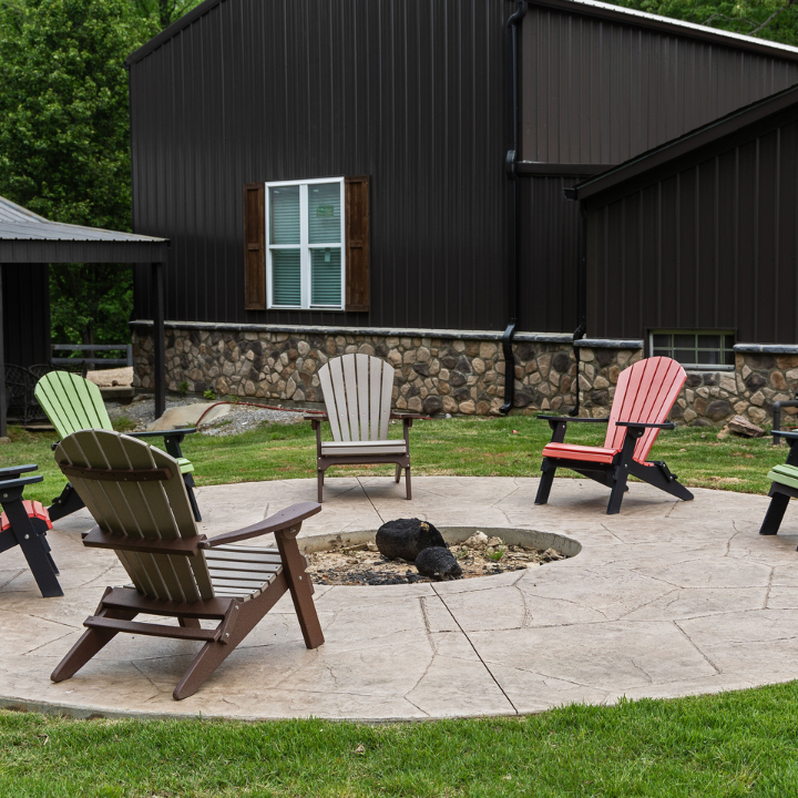 Five Adirondack chairs of various colors arranged in a circle around a stone fire pit on a patio near a dark metal building.