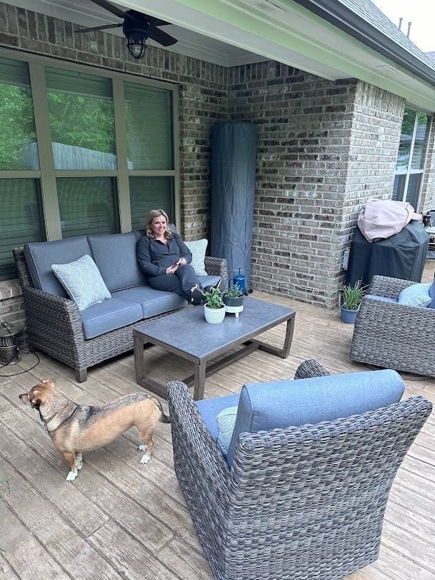 A person sits on a grey outdoor couch on a patio, with a dog standing nearby and a separate grey armchair in the foreground.