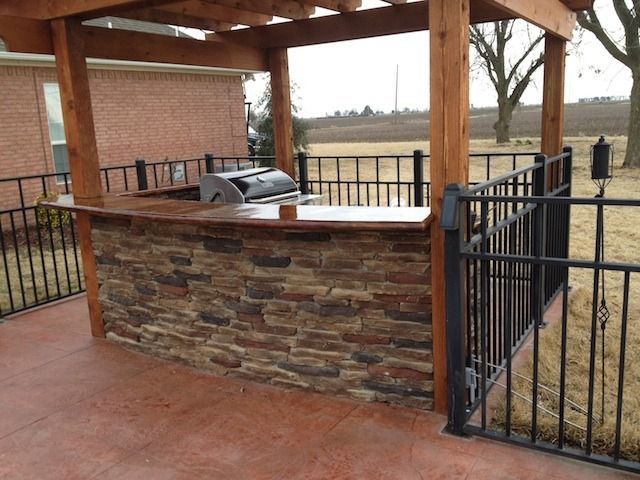 Outdoor stone bar with a grill under a wooden pergola on a patio, surrounded by a black metal railing.