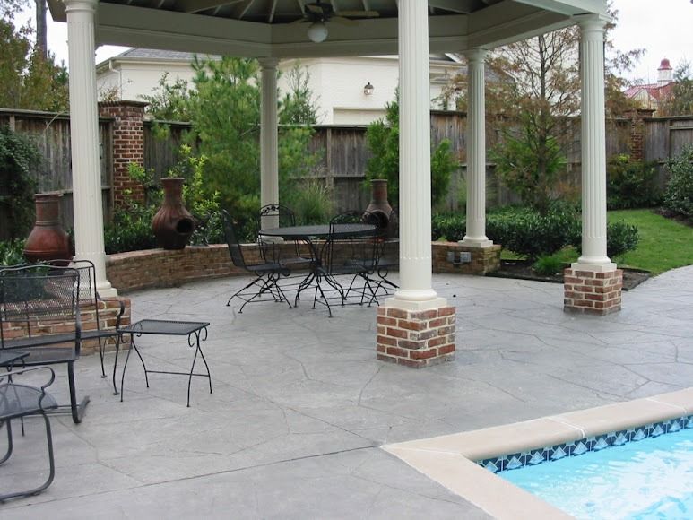 A gazebo with a round table and chairs sits on a concrete patio beside a swimming pool, with a brick wall in the background.