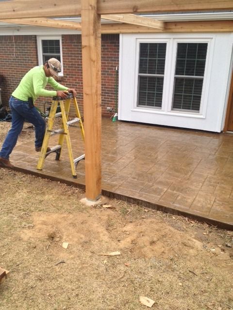 A person in a bright green shirt climbs a yellow ladder on a newly poured, stamped concrete patio by a brick house.