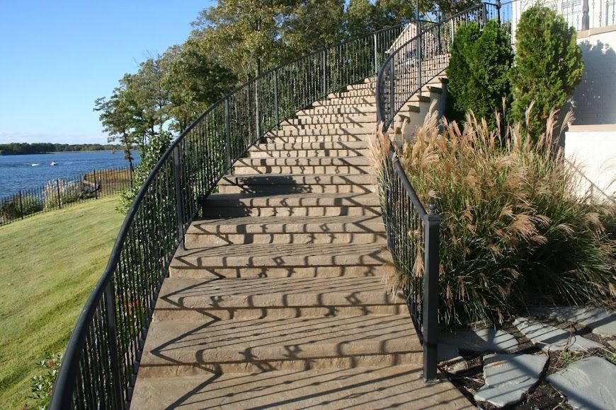 A curved outdoor stone staircase with black metal railings, leading toward a grassy lawn and water under a blue sky.