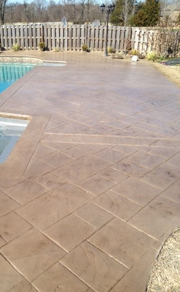 A stamped concrete patio featuring a tan, diagonal stone pattern next to a swimming pool, with a wooden fence in the back.