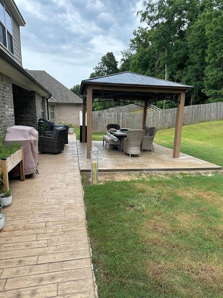 A backyard patio with a covered wooden gazebo, dining furniture, a grill, and a planter box on a stamped concrete path.