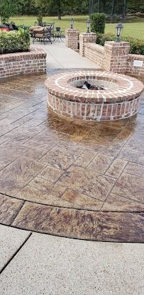 A circular brick fire pit sits on a brown stamped concrete patio, next to a brick retaining wall and a grassy yard.
