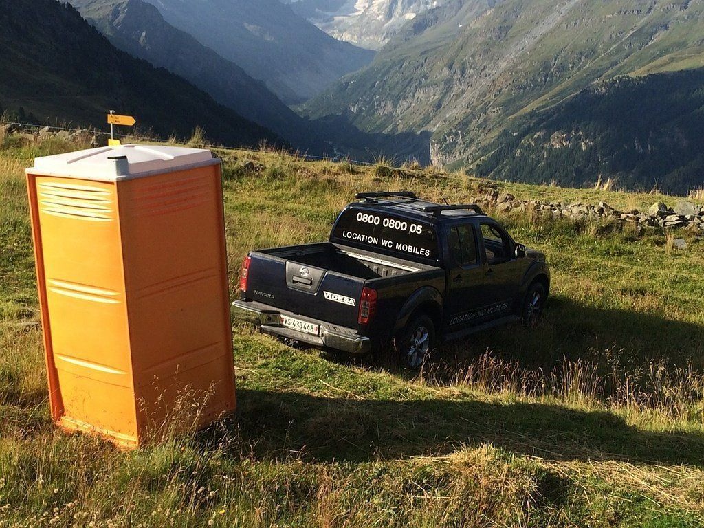 Un camion est garé à côté de toilettes portables dans un champ avec des montagnes en arrière-plan.