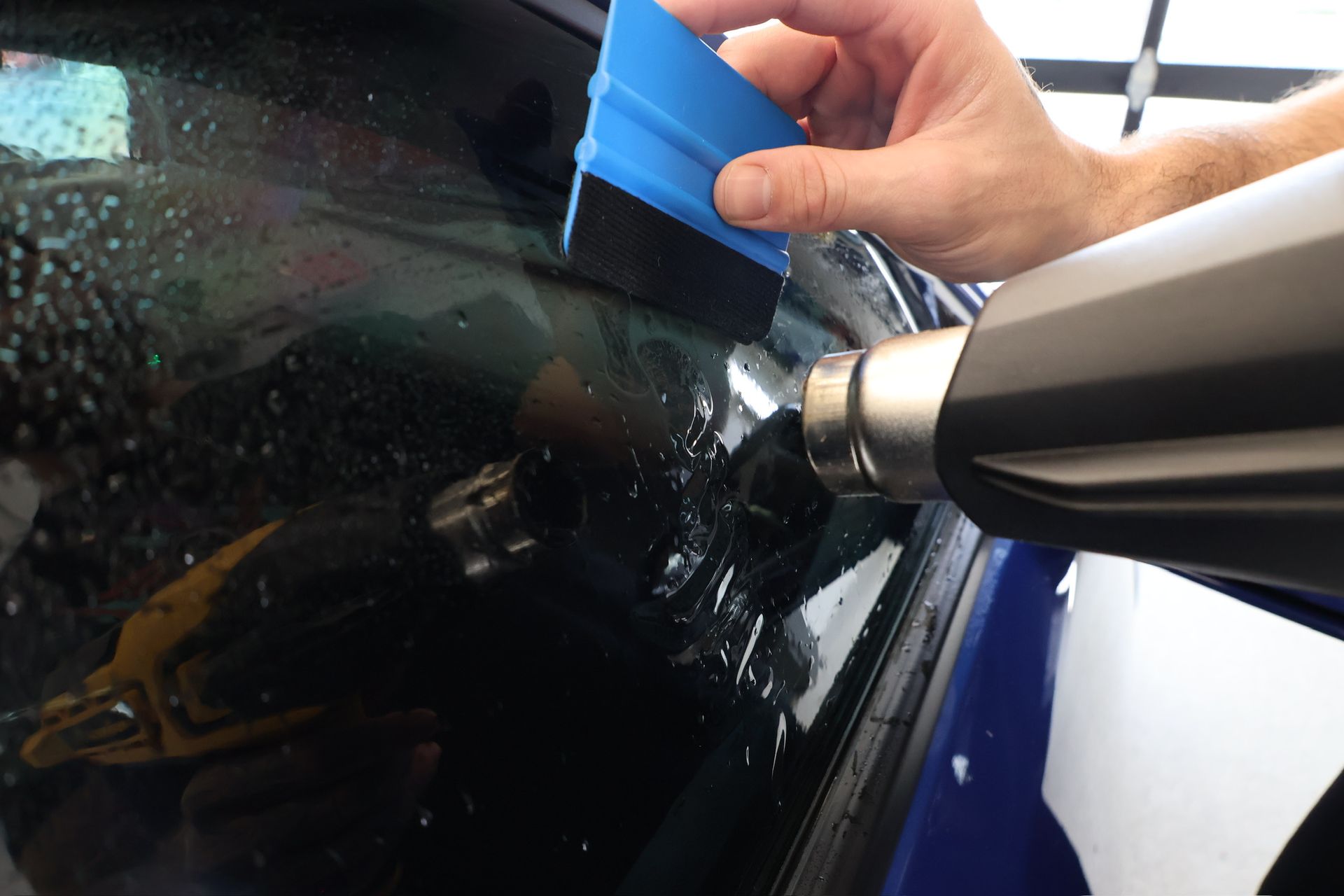 Person using a blue squeegee and heat gun to apply tint to a car window.