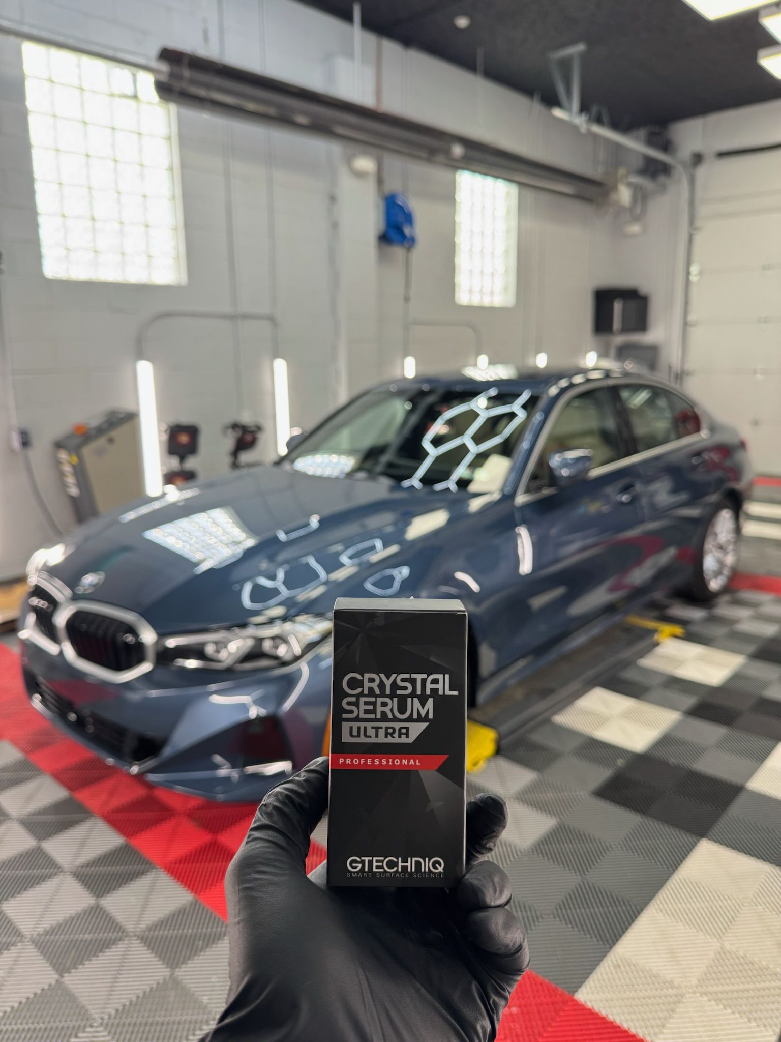 Gloved hand holding a black product box in front of a blue BMW sedan in a garage setting.
