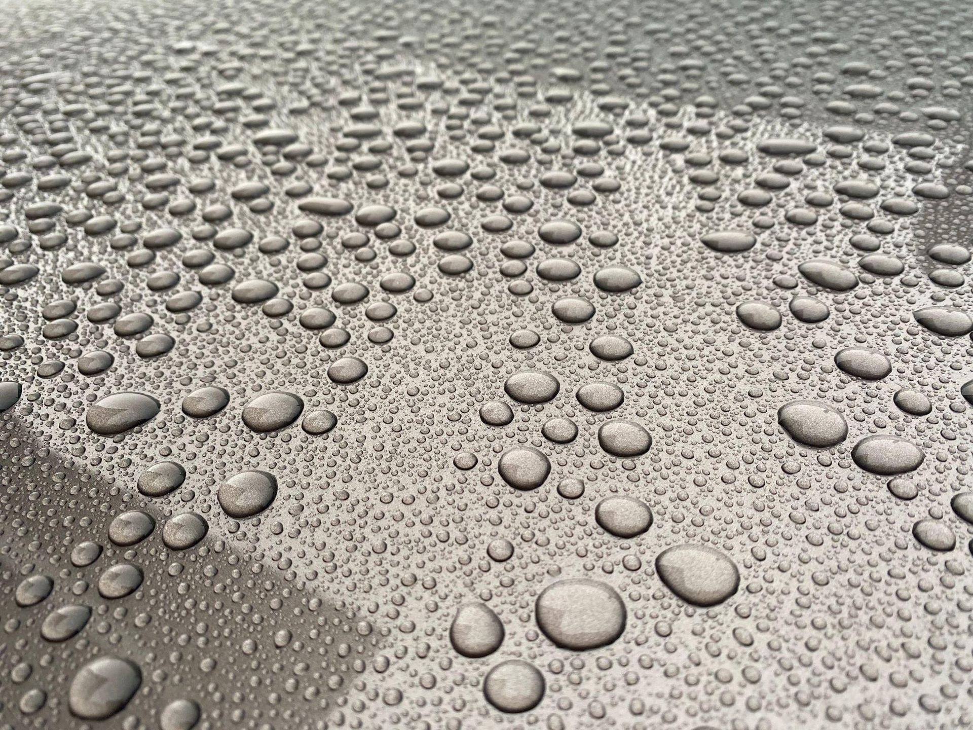 Close-up of a silver car surface covered in many water droplets.