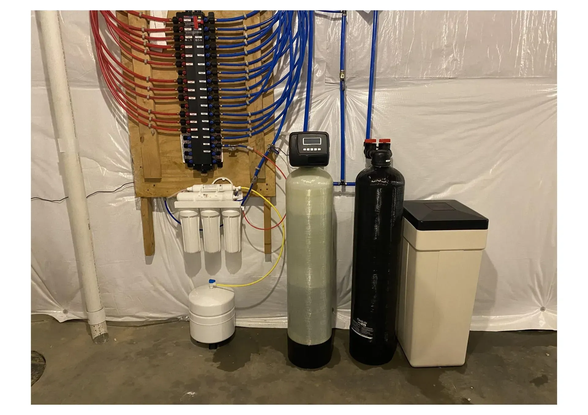 Water treatment system in a basement. Includes a filter, softener, and distribution manifold with colored pipes.