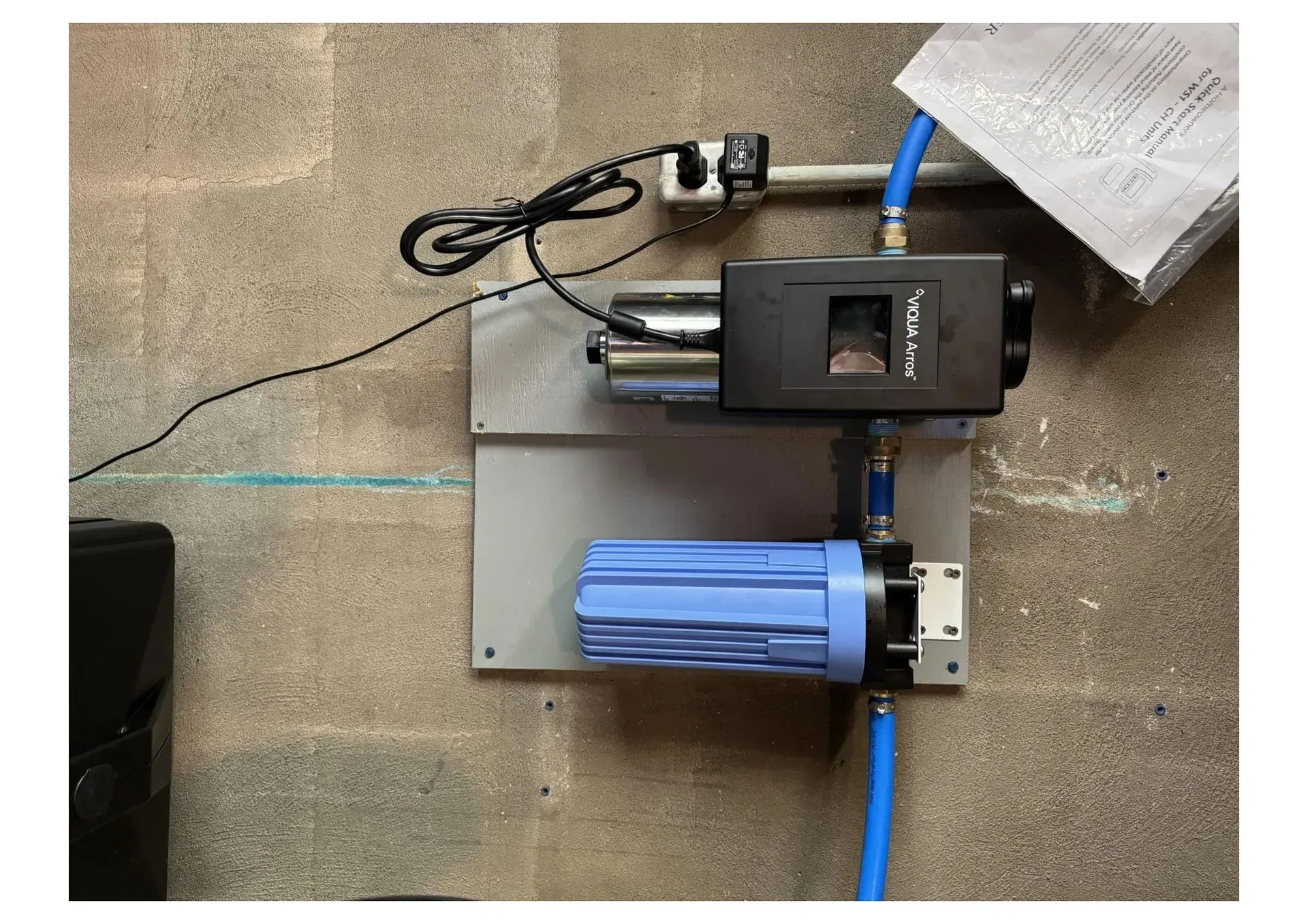 Water filter system mounted on a concrete wall; blue filter, black components, and blue tubing.