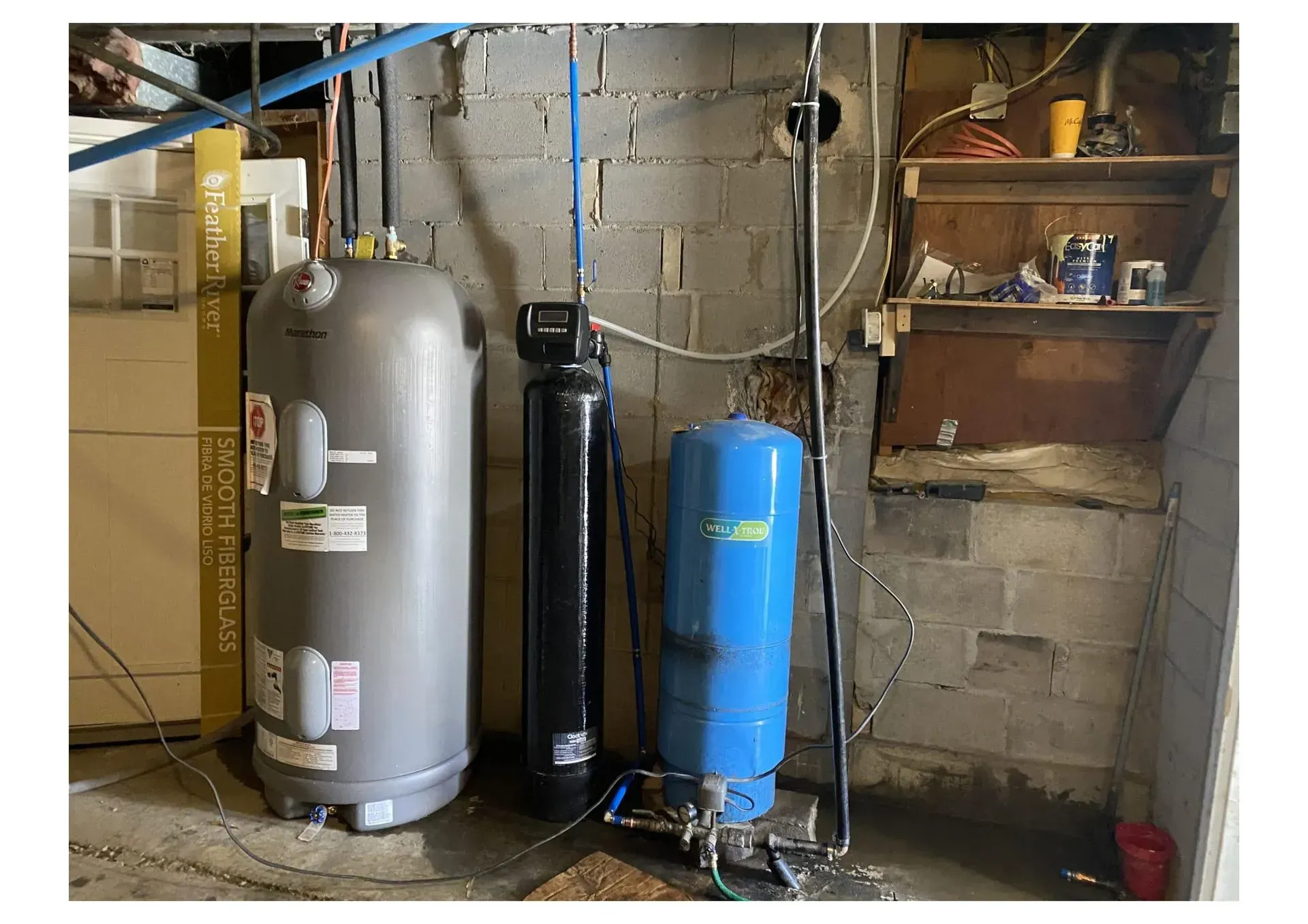 Water heater, water filter, and pressure tank in a basement against a gray block wall.