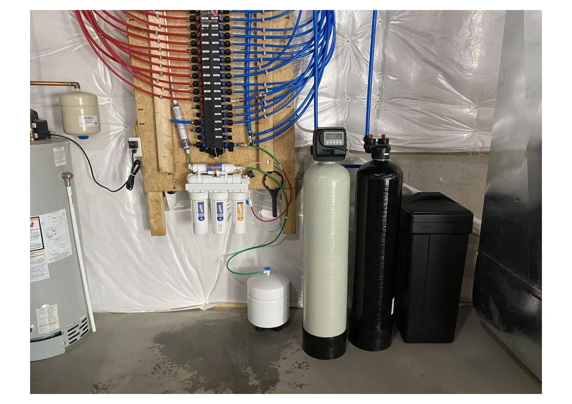 Plumbing and water filtration system in a basement with colored pipes, tanks, and a water heater.