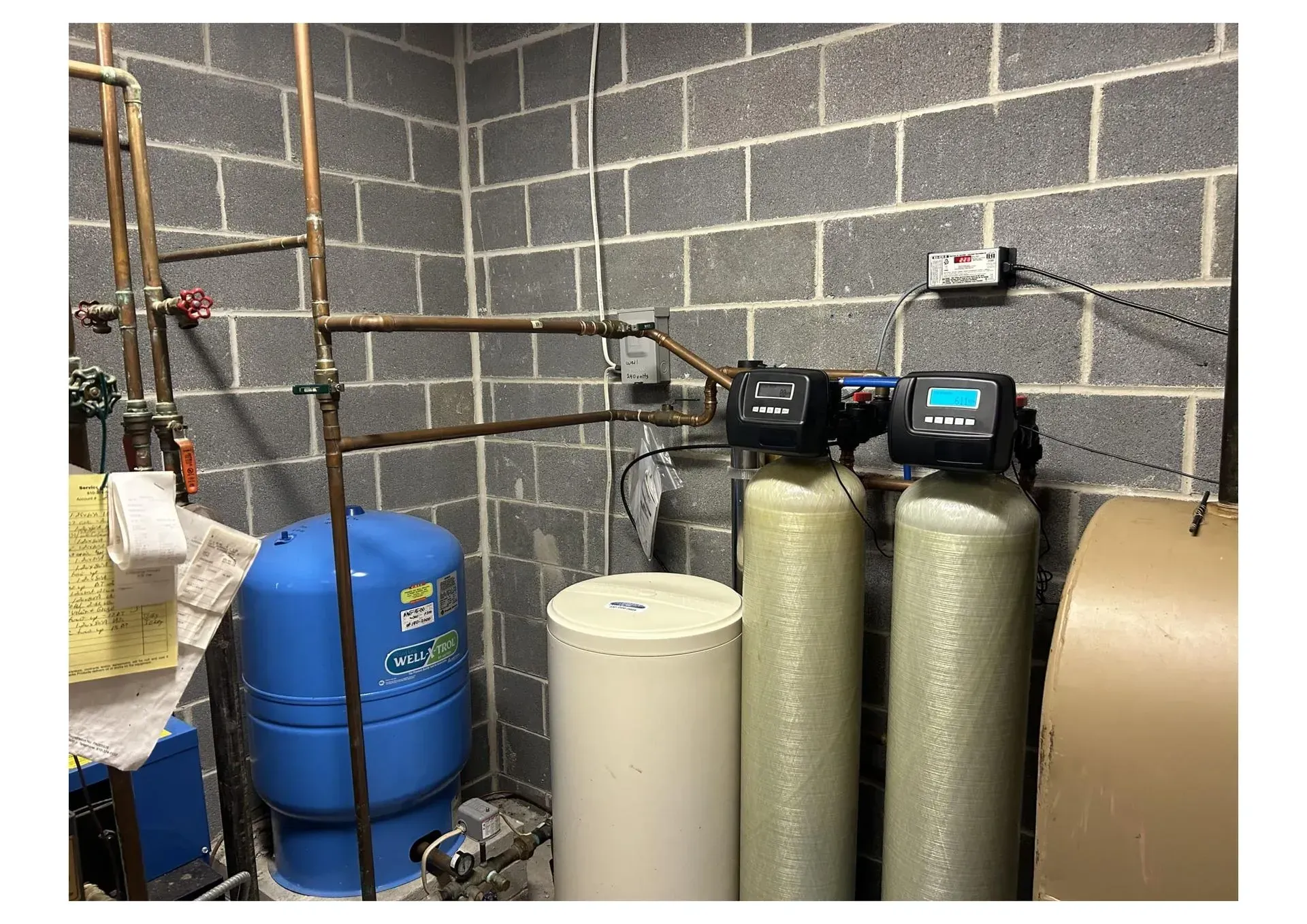 Water filtration and treatment system in a basement setting with gray cinder block walls, pipes, and tanks.