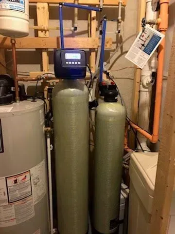 Water softener system with two tall tanks, blue pipes, and digital display, installed near a water heater and utility pipes.