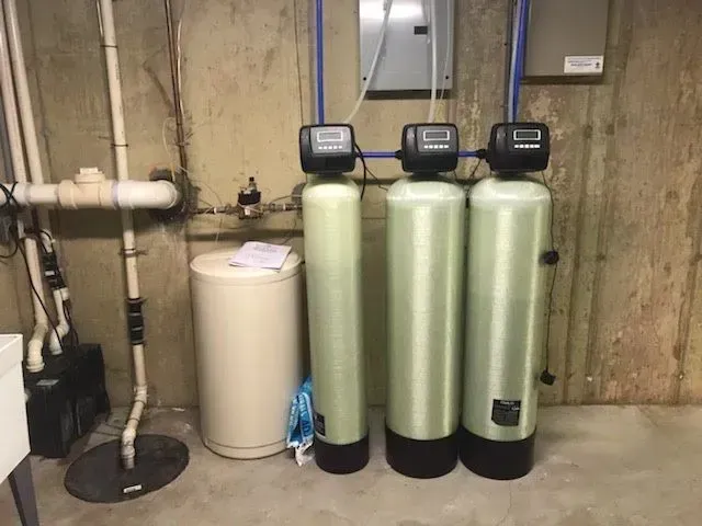 Water treatment system in a basement. Beige tanks and pipes on a concrete floor. Electrical box in the background.