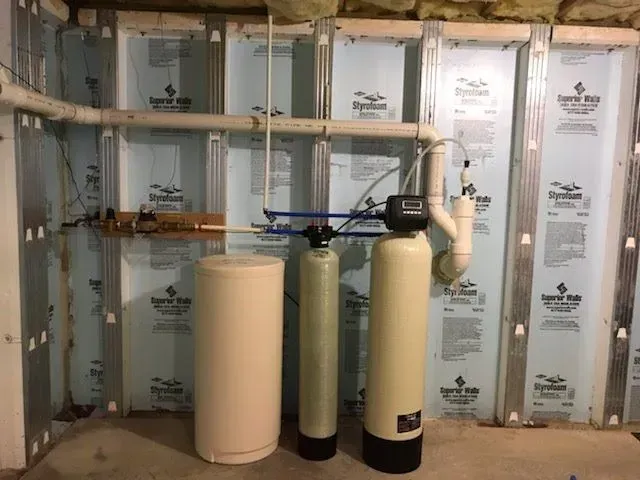 Water treatment system in a basement. Beige tanks and pipes against blue insulation on studs.