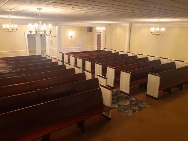 Empty chapel interior with rows of wooden pews, chandeliers, and beige walls.