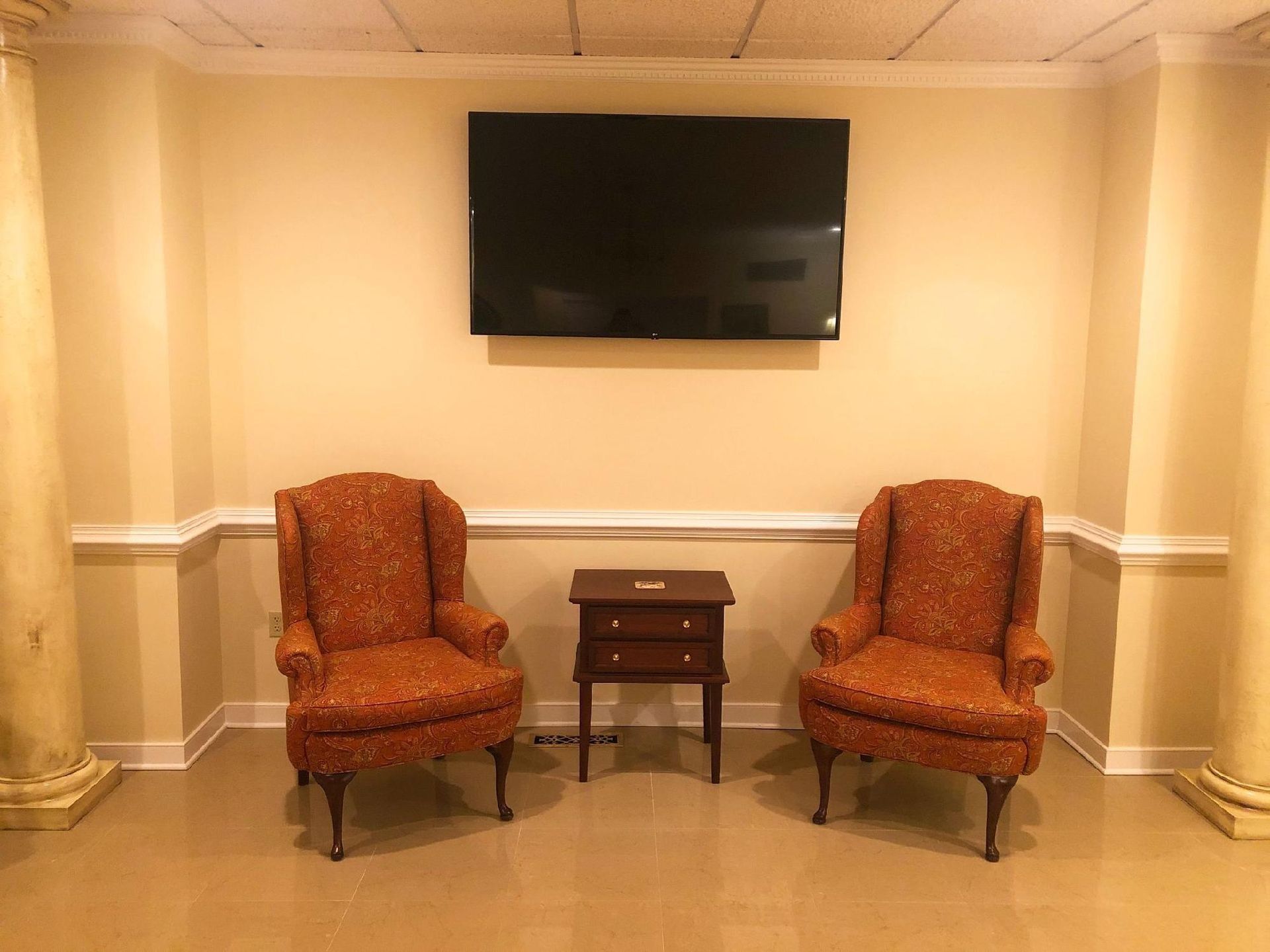 Two patterned armchairs and a small table under a wall-mounted TV in a cream-colored room with columns.