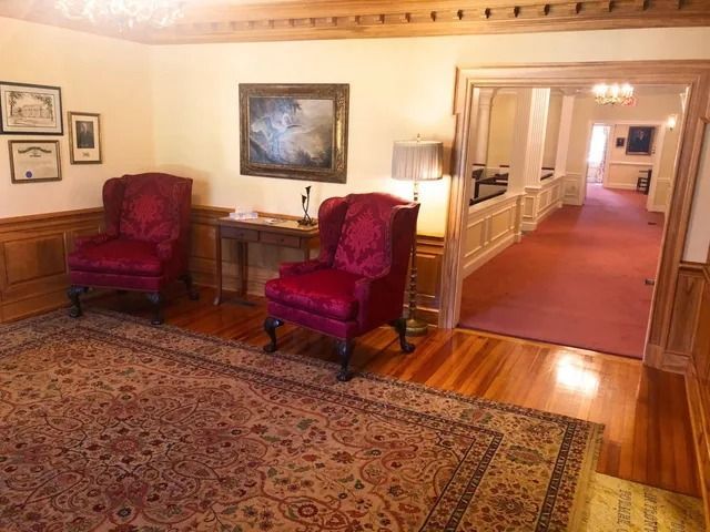 Two red armchairs on patterned rug in a wood-paneled waiting area, doorway to hallway with red carpet.