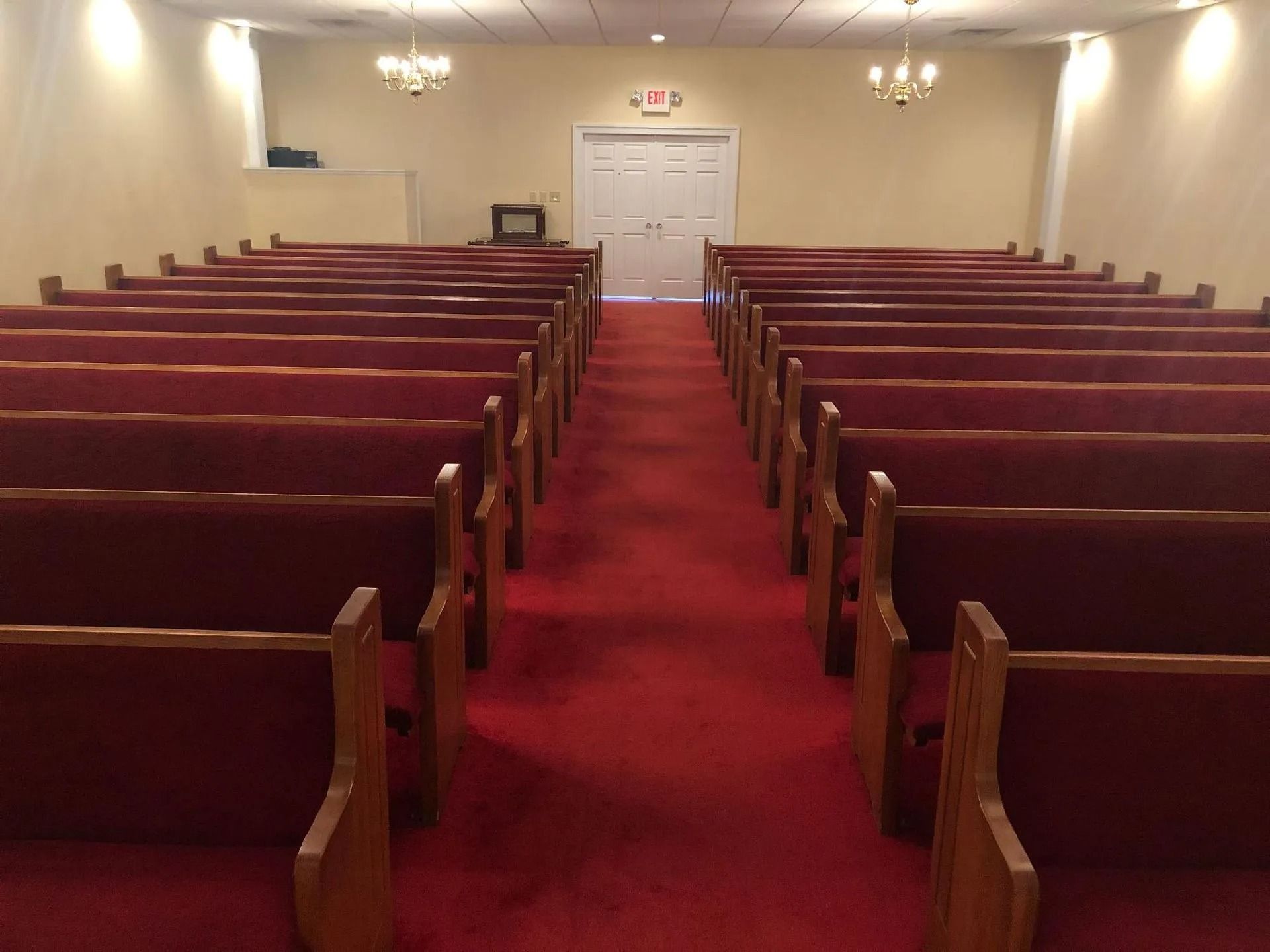 Interior view of a chapel with red carpet, wooden pews, and white walls, facing a closed door.