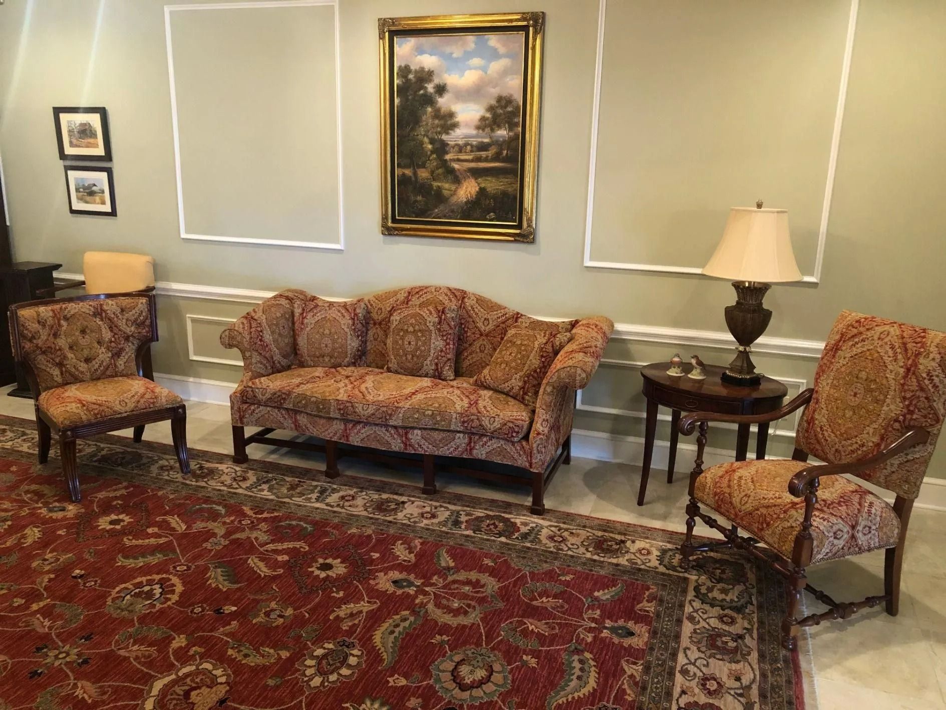 Living room with floral patterned sofa, chairs, and rug, art on the wall, and a lamp on a side table.