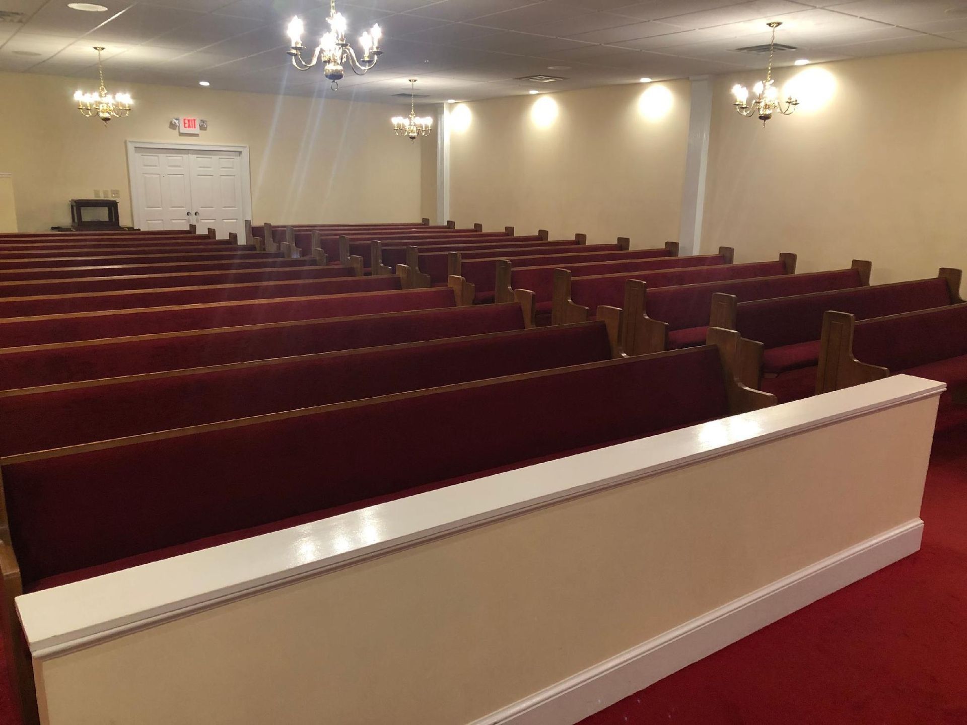 Empty burgundy-seated pews in a cream-walled room with chandeliers and exit sign.