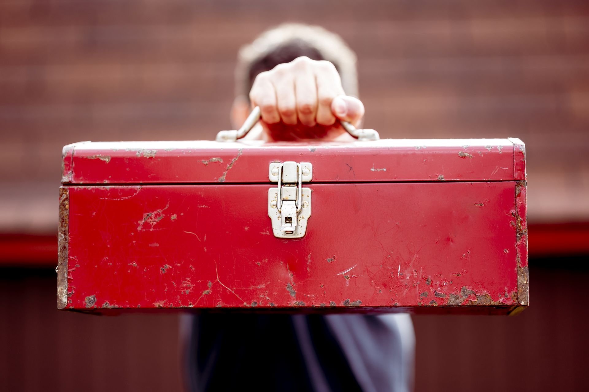 Person holding a weathered, red toolbox towards the viewer.