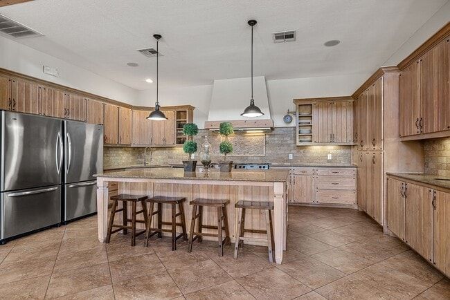 Spacious kitchen with light wood cabinets, stainless steel fridge, island with stools, pendant lights.