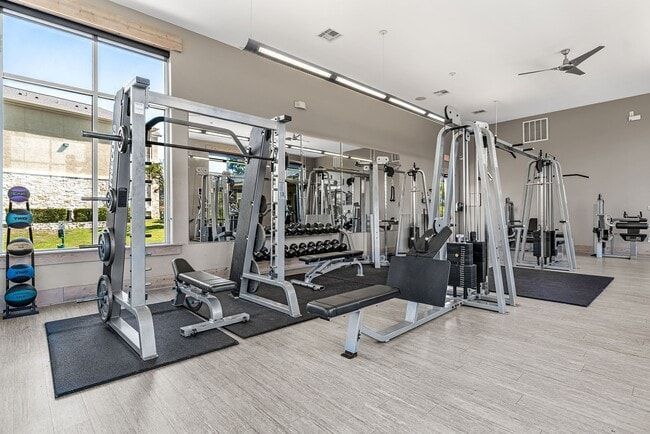 Gym with weight machines, free weights, and a bench, set against a window and mirrored wall.