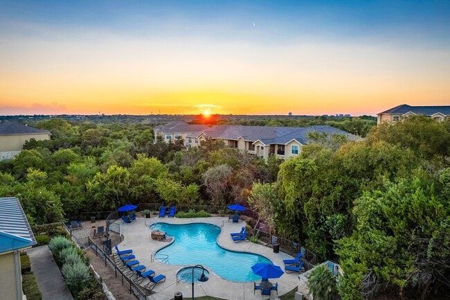 Pool with blue chairs and umbrellas surrounded by trees, buildings, and a sunset.