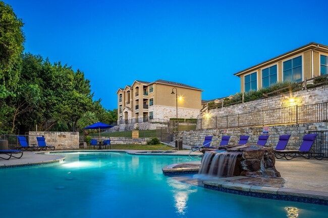 Pool at dusk, with buildings, lounge chairs, waterfall feature, and a blue sky.