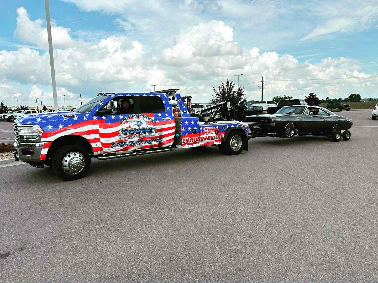 American flag-themed tow truck pulling a classic car on a trailer, parked outdoors under a cloudy sky.