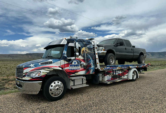 Tow truck with American flag design, hauling a dark pickup truck on a dirt road in a desert landscape.