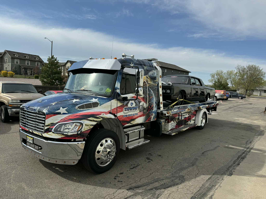 Tow truck with American flag-themed paint hauling a classic car on a sunny day.