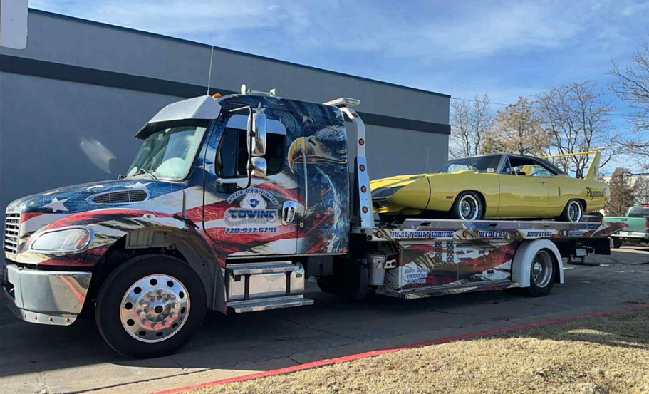 Tow truck with a yellow classic car on its flatbed, parked in front of a building.