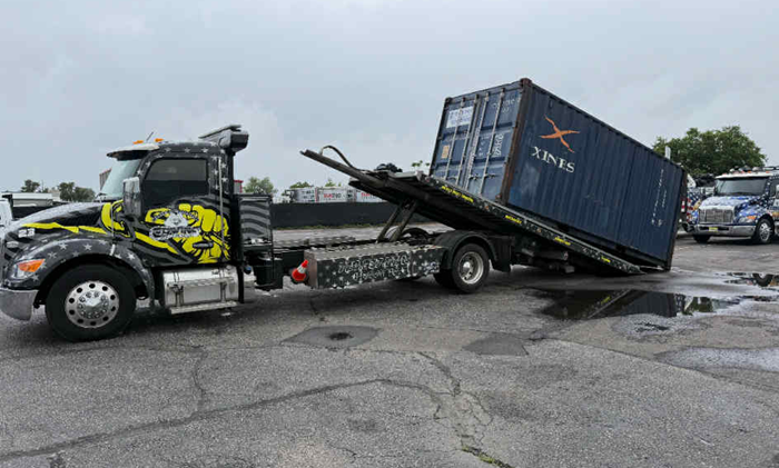 Truck towing a blue shipping container; truck bed tilted. Gray pavement, overcast day.