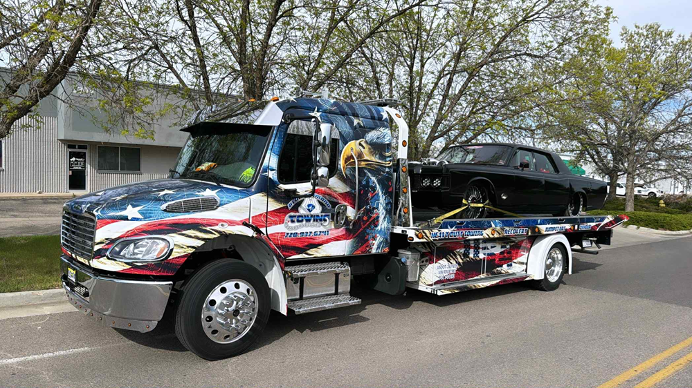 A patriotic tow truck with American flag graphics hauling a black classic car on a sunny day.