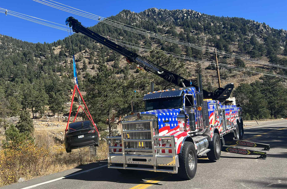 Tow truck lifting a car, roadside near mountains and power lines. Truck has patriotic design.