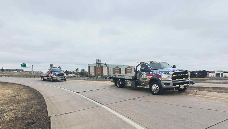 Two tow trucks driving on a highway, gray sky in the background. One truck is in front of the other.